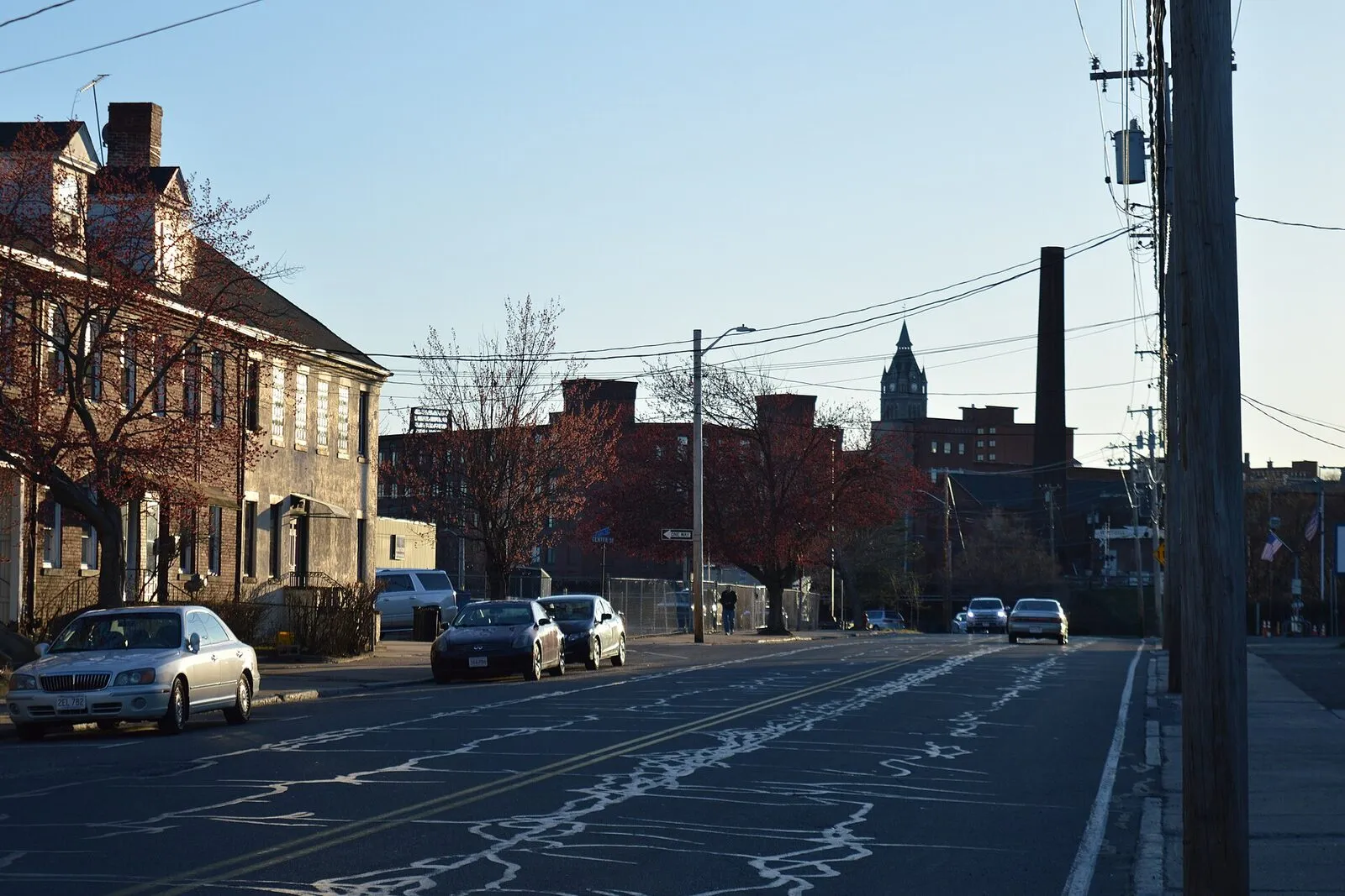 The Holyoke skyline seen from Canal Street in The Flats on an overcast spring afternoon — a foreground row of late-19th-century brick rowhouses on the left, a vacant grass lot stretching across the middle distance, the green-domed clock tower of Holyoke City Hall rising in the center-right of the background, the smaller brick towers of the Lyman Mills / Open Square complex to its left, mature deciduous trees in early leaf along the street, and a flat gray sky overhead.
