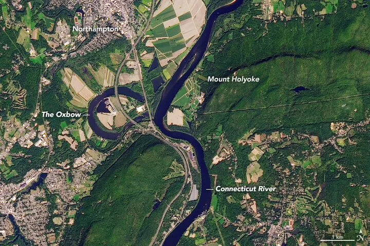 A NASA Landsat satellite image of the Connecticut River Oxbow on the Easthampton–Northampton border in late September — a wide horseshoe of dark blue still water curving back on itself in the bottomland between the green wooded ridge of Mount Tom and the patchwork of farm fields on the river's old course, with the active modern channel of the Connecticut River cutting across the frame and the dark green of the Arcadia Wildlife Sanctuary's floodplain forest hugging the oxbow's edge.