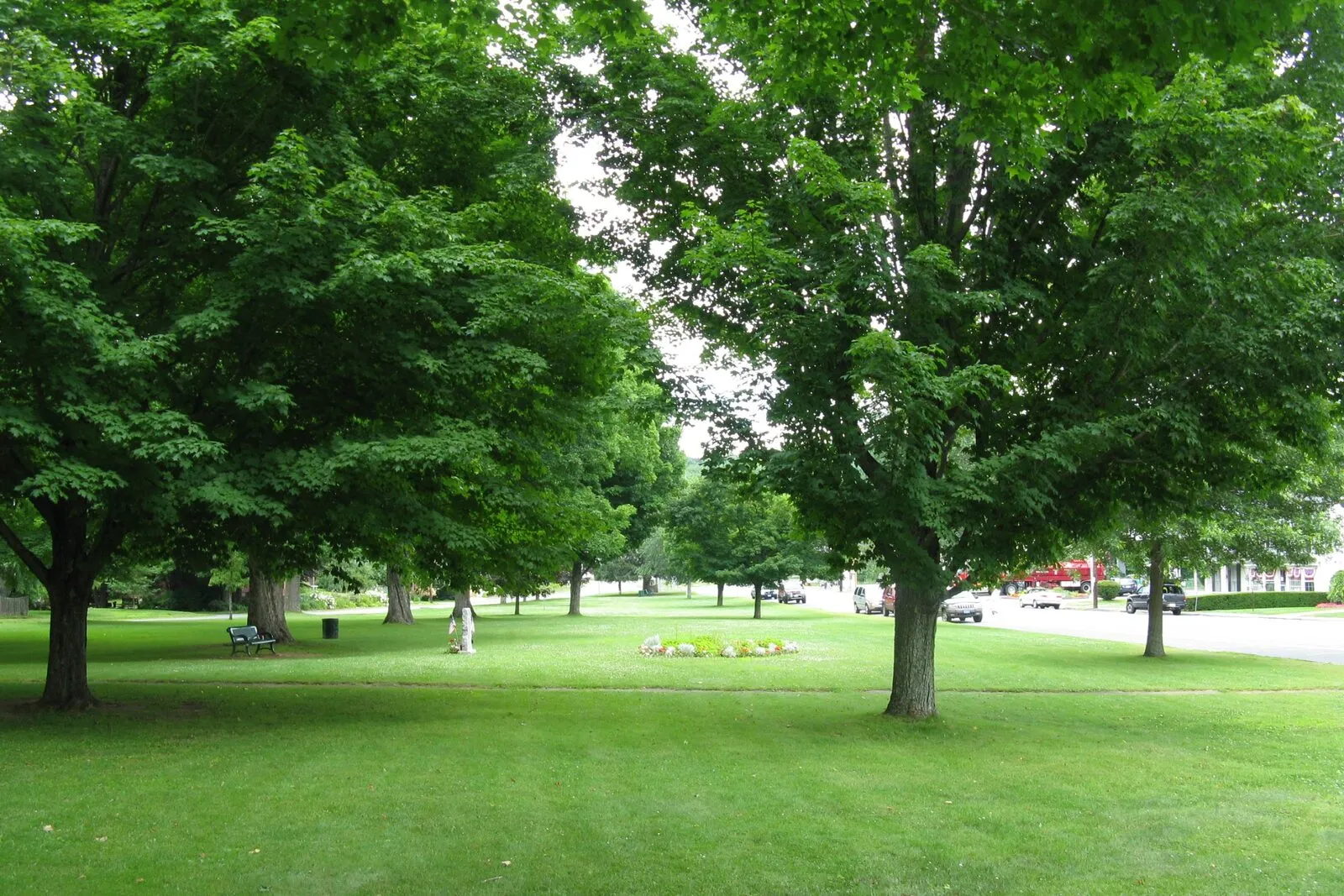 The Brimfield town common on a sunny summer afternoon: a long, narrow grassy green flanked by paved roads, with mature deciduous trees in full leaf casting shadows across the lawn, a tall white-clapboard New England meetinghouse with a multi-stage square bell-tower and lantern cupola standing prominently at the far end, smaller white-painted civic buildings and houses lining the sides of the common, a flagpole at center, low stone curbing along the edges, and a deep-blue sky with thin scattered clouds overhead.