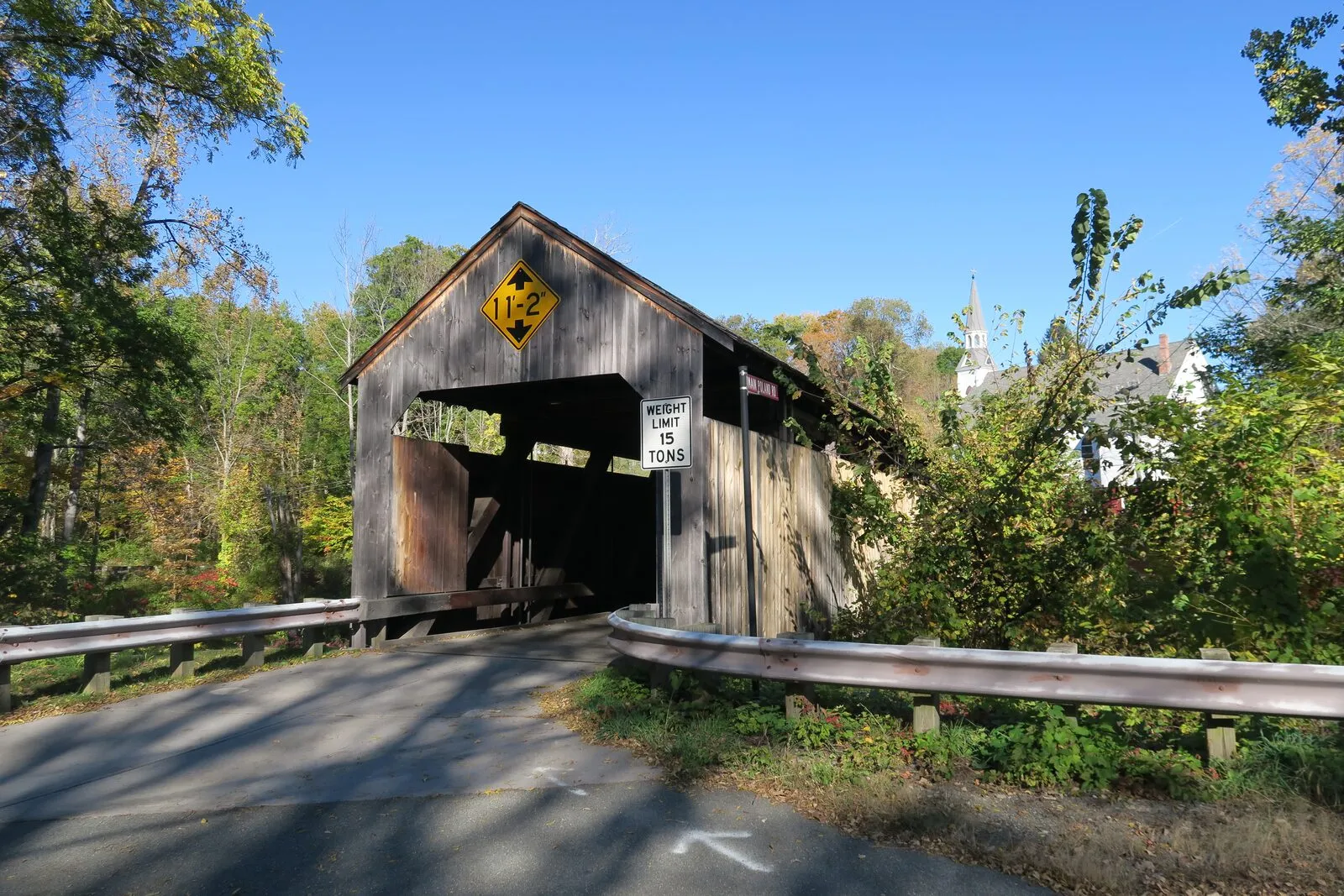 The Burkeville Covered Bridge from the road approach on a sunny autumn afternoon. A weathered grey board-and-batten covered bridge with a low gabled roof crossing over the South River, a yellow weight-limit sign mounted above the dark portal, a 'WEIGHT LIMIT 4 TONS' sign on the right portal post, metal guardrails leading into the bridge entrance, deciduous trees with green and yellow foliage on the left, and a slim white church spire poking above the trees on the right under a clear blue sky.