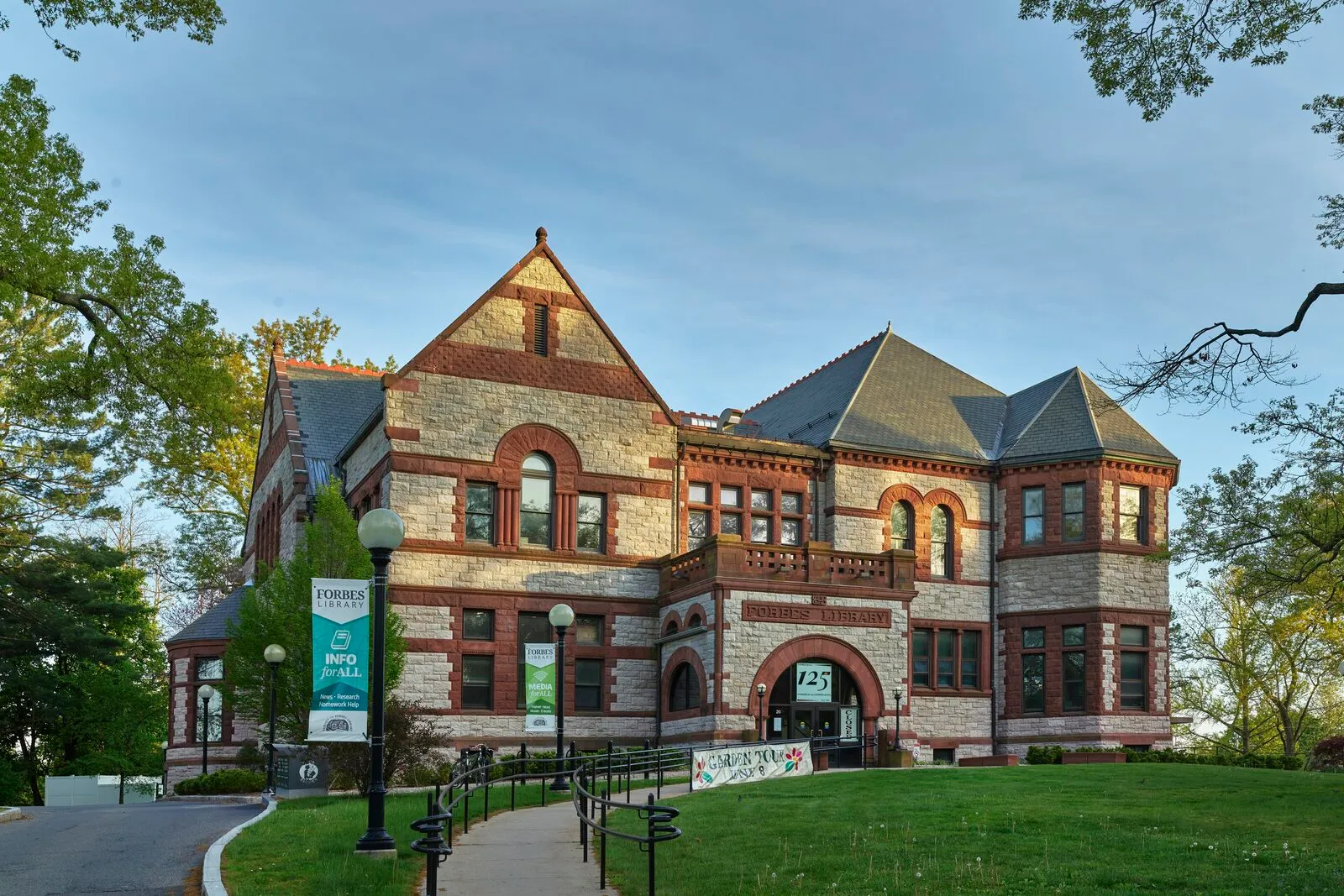 The 1894 Forbes Library in Northampton on a clear late-spring afternoon. A Richardsonian Romanesque public library built of rough-faced two-tone sandstone, with bands of round-arched windows along the lower floor, paired square windows above, a tall central gable flanked by stepped slate roofs, a recessed arched entrance reached by a ramped walkway, mature deciduous trees with new leaves framing the right side, and a green lawn under a blue sky with light cumulus.