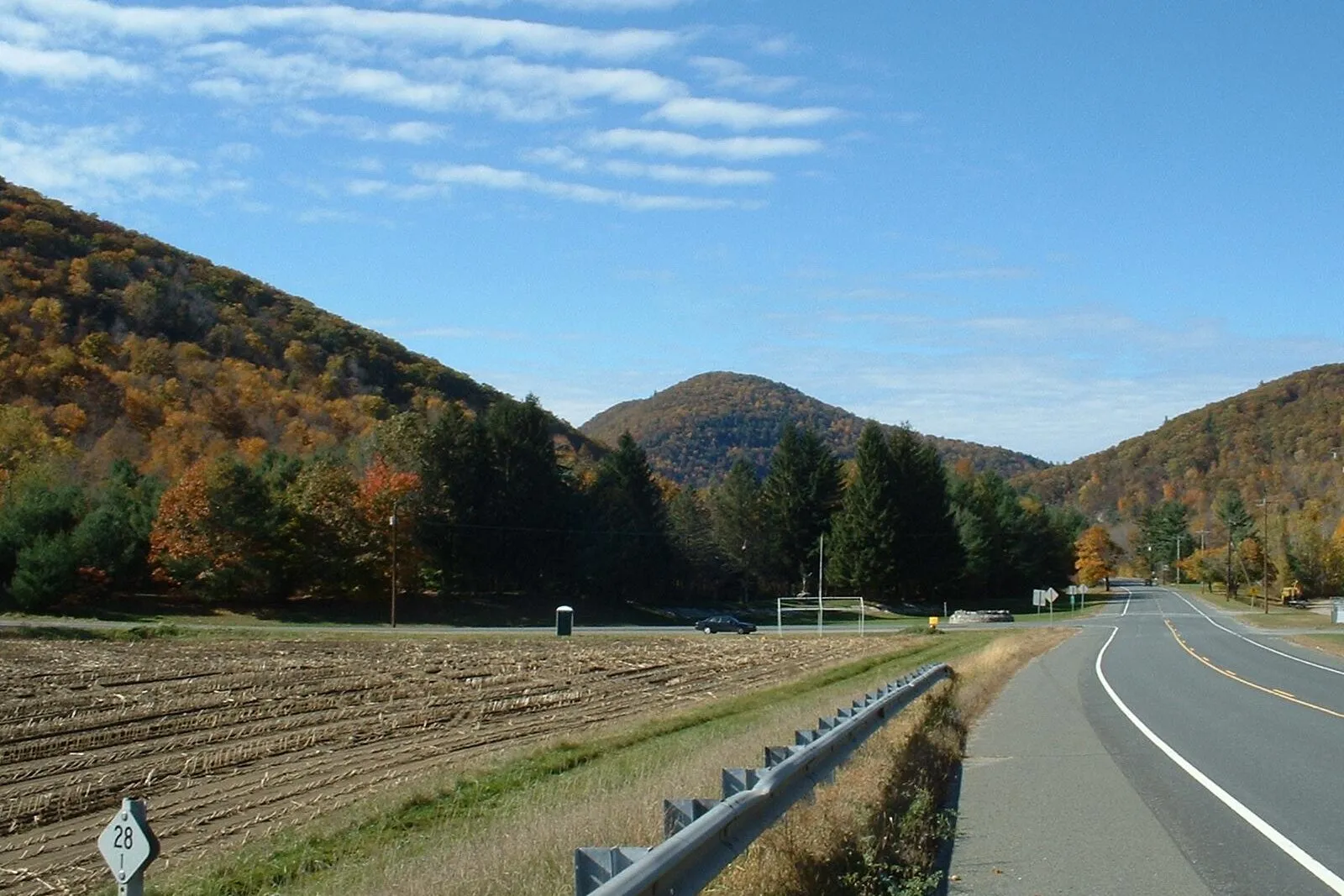 Looking west along Route 2 — the Mohawk Trail — through the Deerfield River valley in Charlemont on a clear, sunny fall day, with peak orange-and-yellow foliage covering the steep flanking ridges of two layered mountains, a third smaller wooded peak rising in the gap between them, dark-green conifers in a midground hedgerow, a brown harvested cornfield with a milepost-28 marker in the left foreground, and the empty two-lane highway curving to the right along a guardrail under a deep-blue sky scattered with thin cirrus clouds.