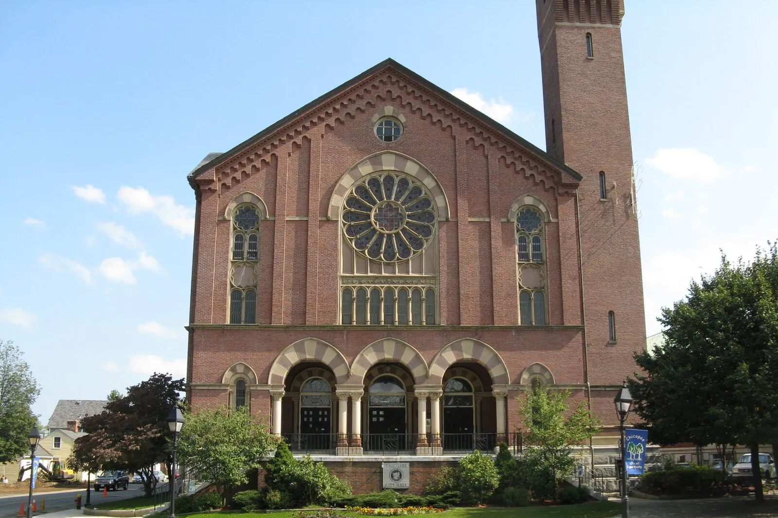 Chicopee City Hall on a sunny late-summer afternoon: a tall red-brick 1871 Romanesque-Revival building on a downtown corner, large central round-arched entry portico with three arches, a big rose window above the entry, twin pointed-arch windows flanking it on either side, a tall square brick clock tower rising on the right side beyond the gable, mature street trees and a low stone wall bordering the sidewalk in the foreground, and a clear blue sky overhead.