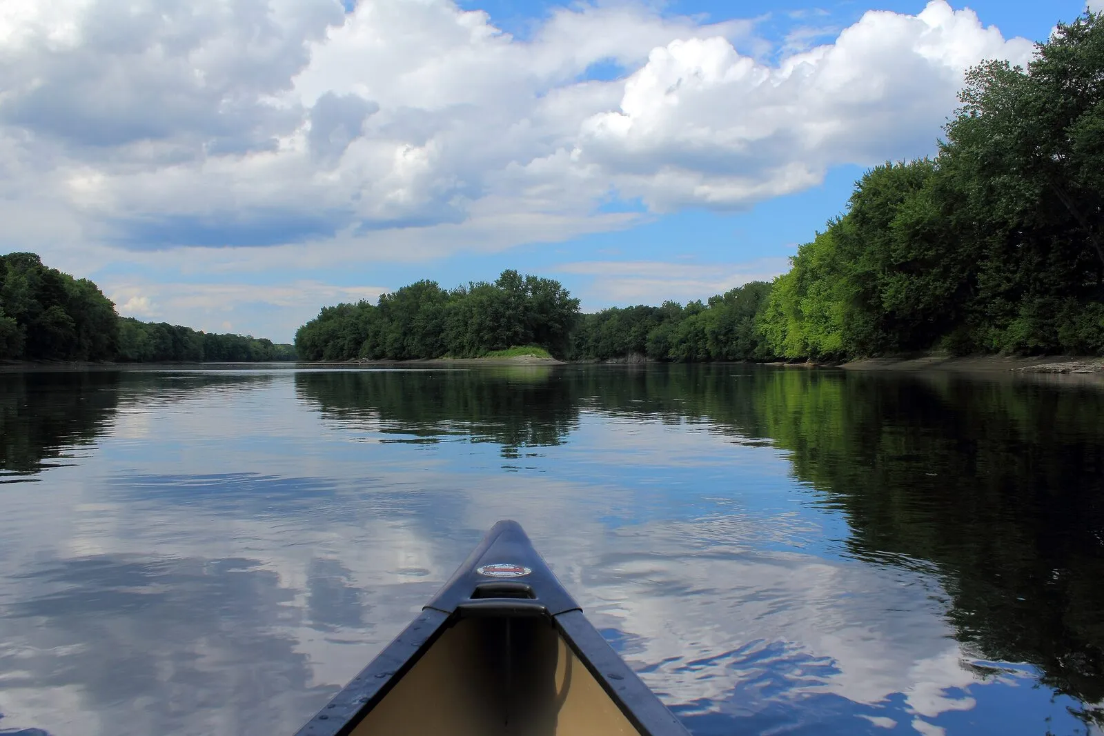 A canoe bow pointing north up a glassy stretch of the Connecticut River near Sunderland on a summer afternoon: dark forested banks reflecting in the still water on either side, a slim sandbar showing in the middle distance, and a deep blue sky with bright white cumulus clouds reflected back from the water in long ripples in front of the canoe.