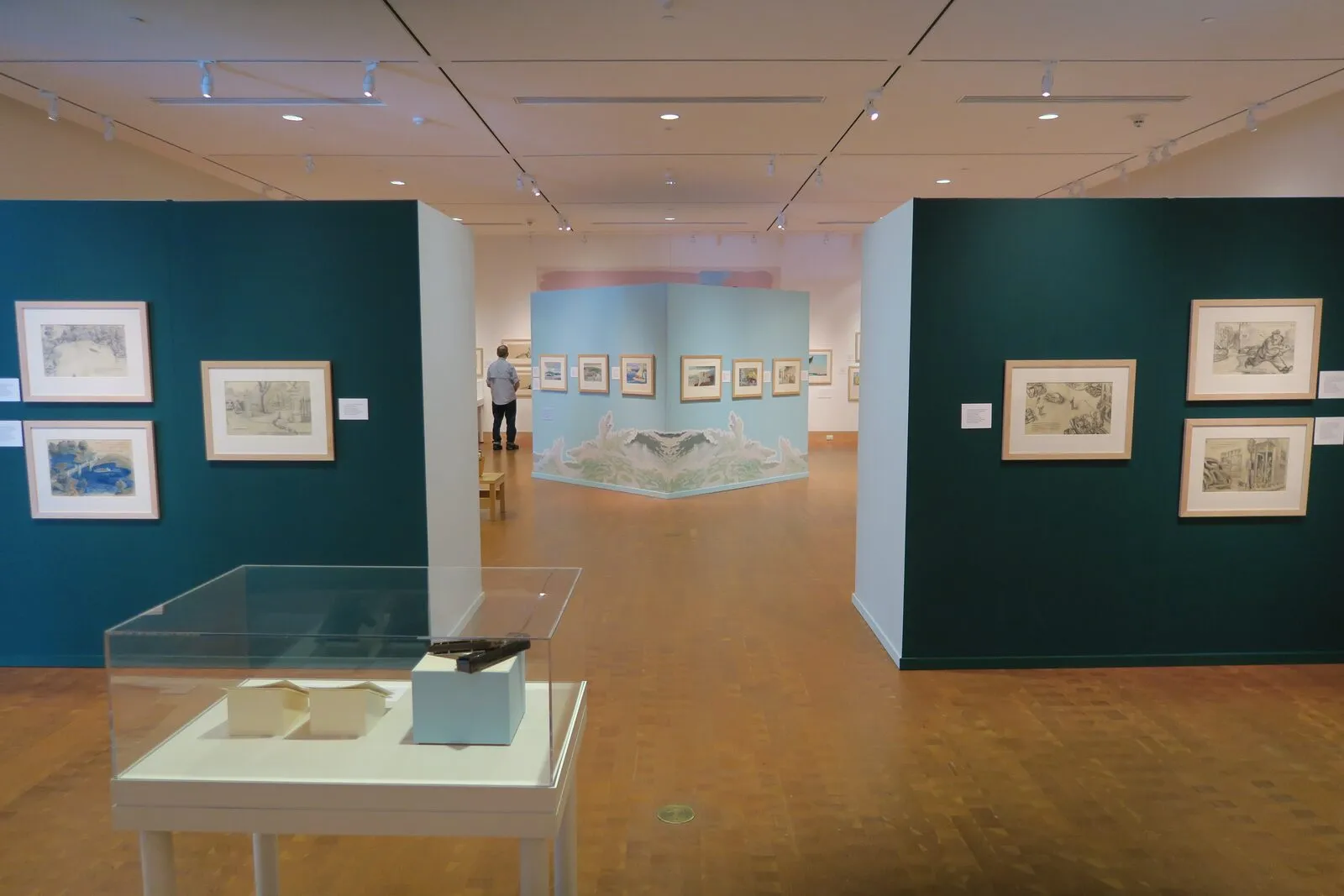 A gallery inside the Eric Carle Museum of Picture Book Art — teal walls hung with framed picture-book illustrations, a sky-blue partition with a painted ocean-wave cutout at its base, a glass display case of small paper book mockups in the foreground, and a warm parquet-wood floor receding into the room.