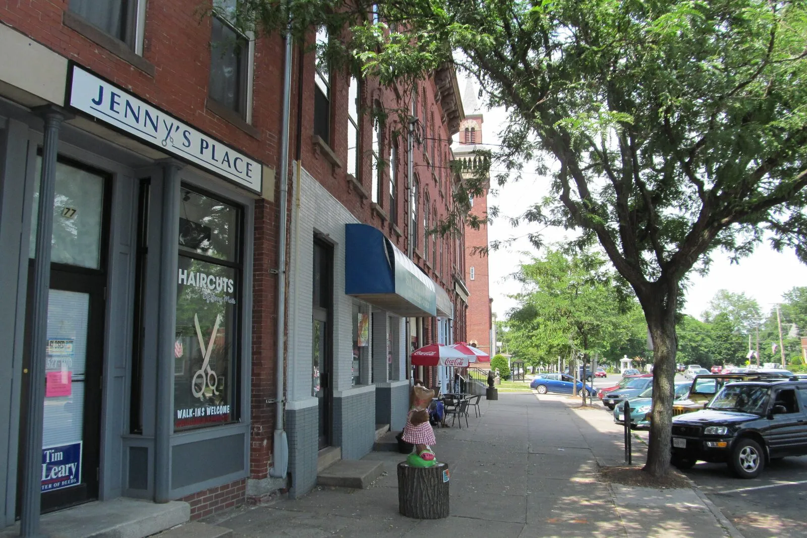 A sidewalk view down Main Street in downtown Easthampton on a summer afternoon — a red-brick Italianate storefront block with Jenny's Place hair salon at the near corner, a red café umbrella further down, a distinctive mansard-towered building in the middle distance, a leafy street tree on the right, and parked cars along the curb.