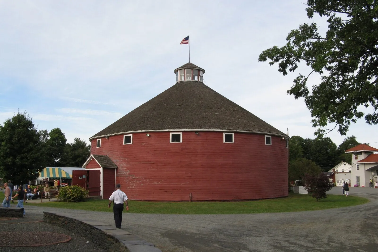 The Roundhouse at the Franklin County Fairgrounds in Greenfield on a partly cloudy late-summer afternoon: a large circular barn painted dark red with a low conical shingled roof topped by a small white cupola flying a US flag, a row of small white-trimmed square windows set just below the eaves, a smaller dark-red shed wing attached on the left side, a green-and-white striped awning in shade at far left with several people seated beneath it at picnic tables, a man in a white short-sleeved shirt and dark trousers walking along a flagstone path in the foreground, a paved drive curving across the front of the barn, mature deciduous trees flanking the building, and the corner of a pale-yellow outbuilding visible at the right edge.