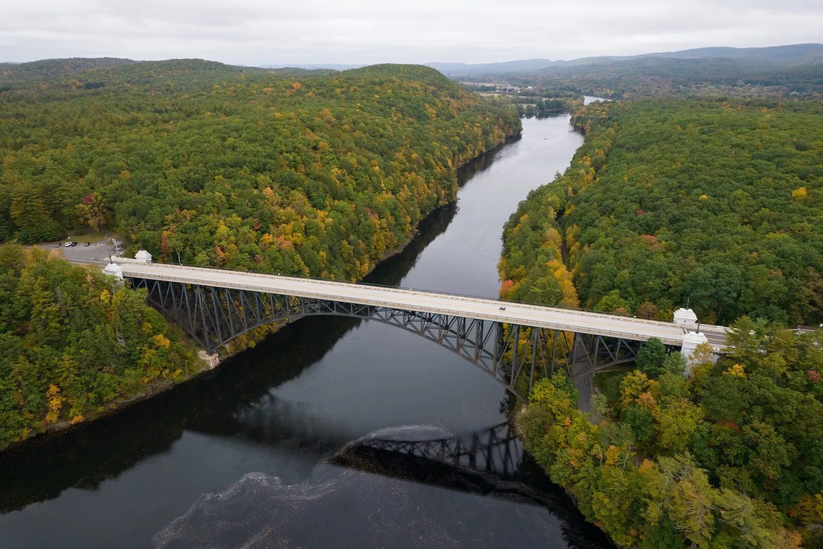 The French King Bridge seen from the south on an overcast October afternoon: a long three-span steel cantilever-arch bridge in dull green-gray paint crossing the Connecticut River high above the water, the steel arches springing from each wooded bank and meeting in the middle, dense fall foliage on both sides in mixed orange, yellow, and still-green hardwoods, the river running calm and dark beneath the span and continuing north toward distant low hills, a small cluster of buildings just visible at the far end of the river under a flat pale-gray sky.