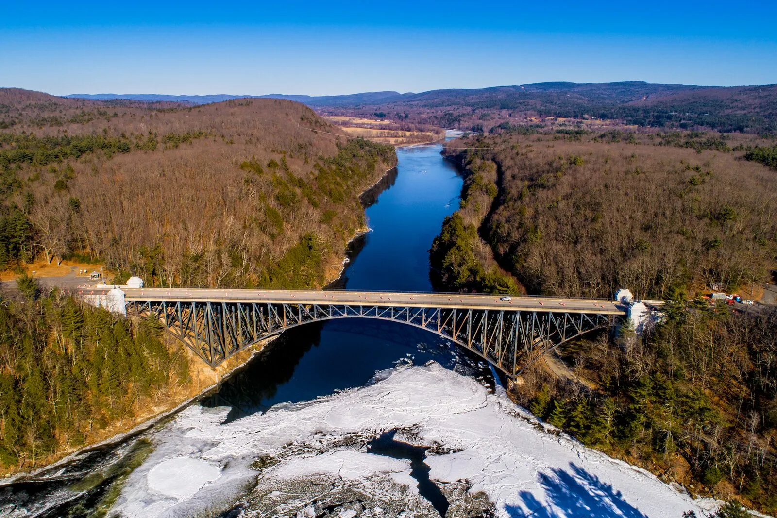 Aerial drone view of the French King Bridge over the Connecticut River between Gill and Erving on a clear winter day. A long three-span steel cantilever-arch bridge in dull green-gray paint crossing high above the river, the river itself partly frozen with broad sheets of pale-gray ice and patches of dark open water curving through the gorge below the bridge, dark wooded hills on both banks in bare-branch winter brown, the river continuing north into the distance under a clear pale-blue sky, and small specks of cars on the bridge deck giving a sense of scale.