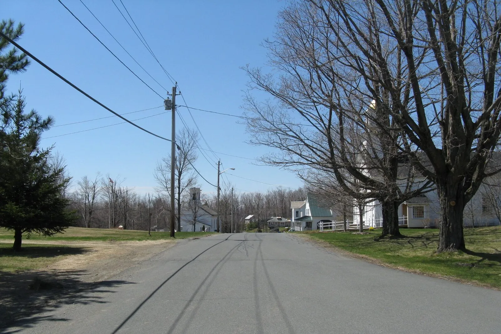 Looking east along East Main Street through Heath Center on a bright, clear early-spring afternoon. The narrow paved village road runs straight ahead between mown grass shoulders, a small white-clapboard 19th-century church with a square belfry sits set back at midground on the left, a much larger white-clapboard meetinghouse-style building rises behind a row of bare deciduous trees on the right, scattered single-story white houses are arranged along the south side of the road, a low utility pole with a streetlight stands in the middle distance, dark-green conifers crowd the left foreground, and a deep cloudless blue sky stretches above the leafless tree limbs.