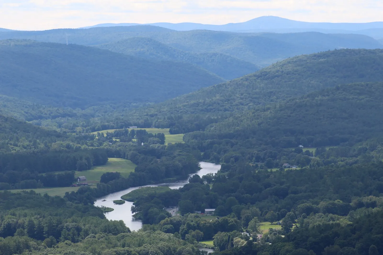 A wide view from a high overlook looks west across rolling, forested hills under a hazy summer sky. In the middle distance, a meandering river winds through a green valley dotted with farm fields, a few houses, and a small white-steepled church. Layers of forested ridges recede into the distance, fading from deep green to a soft blue-grey. On the far horizon, a single rounded summit with a small tower stands above the surrounding hills — Mount Greylock. No people are visible.