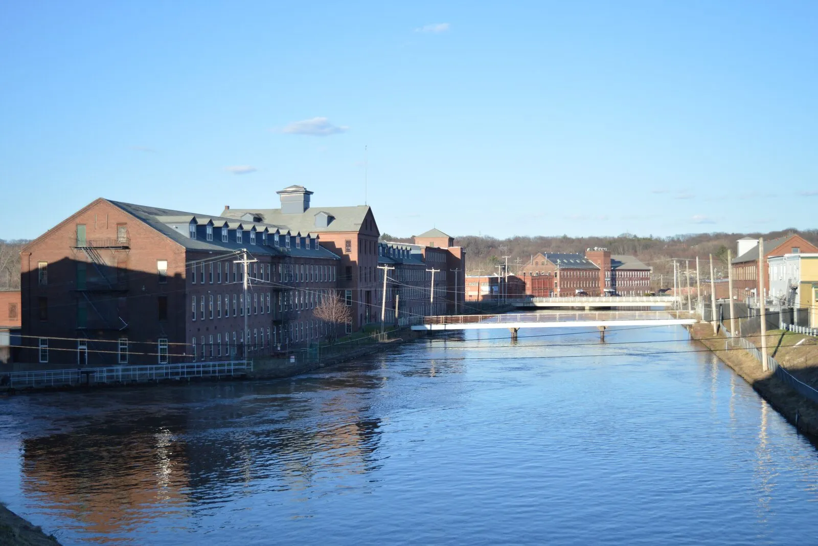 The Second Level Canal in downtown Holyoke on a spring afternoon: a long red-brick mill building (the former American Thread Company) on the left bank, a pedestrian footbridge crossing midway, more mill buildings on the far side, and a wooded ridge rising behind under a clear blue sky.