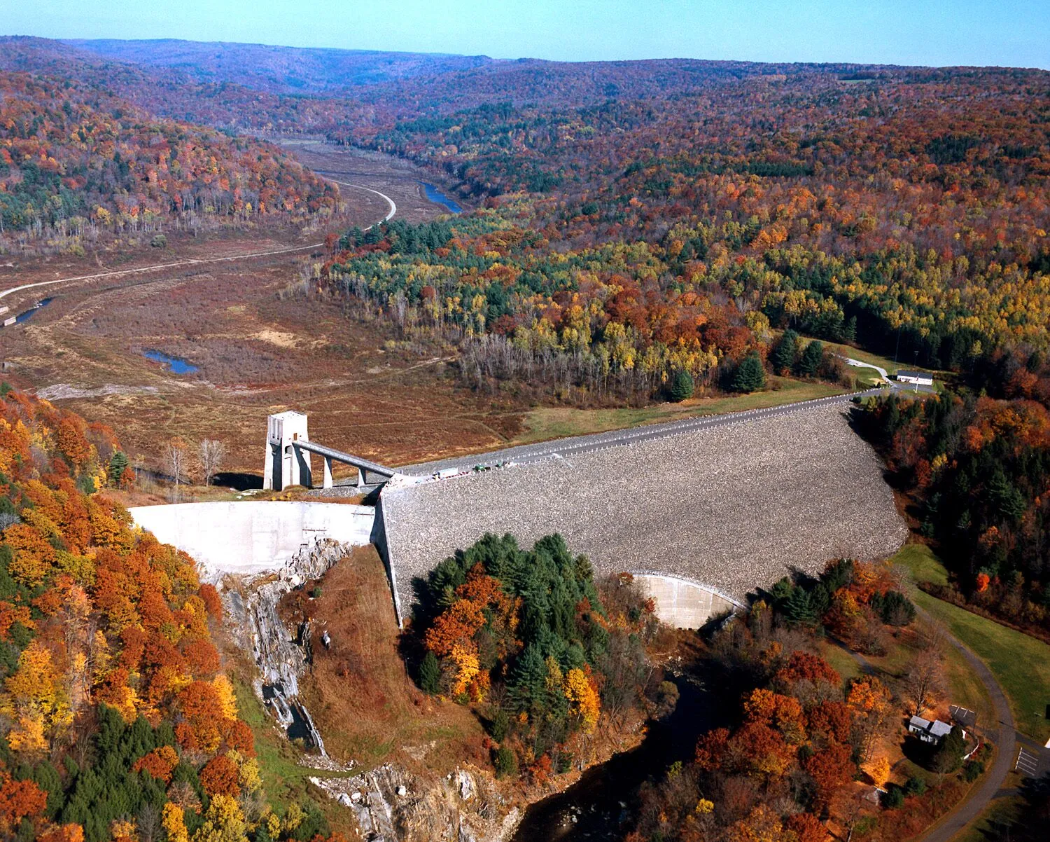 Aerial view of Knightville Dam in autumn — a long earthen embankment runs diagonally across a forested valley, holding back the East Branch of the Westfield River. The dam face is a gently sloped wall of grass and rock; a paved access road traces its crest. Below the dam, the river resumes its winding course through bare-trunked floodplain woods. The surrounding hills are covered in patchy yellow, orange, and brown fall foliage with stretches of dark evergreen, and a low blue ridgeline rises behind the structure.