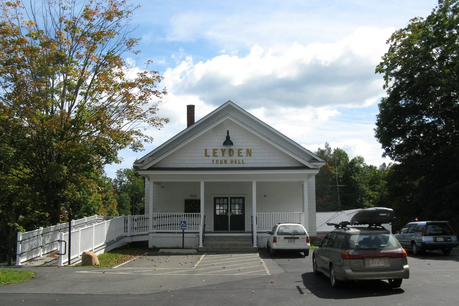 Leyden Town Hall on a bright early-September afternoon — a modest single-story white-clapboard civic building with a shallow pedimented gable end facing the small paved parking lot, the words LEYDEN TOWN HALL painted in dark lettering across the frieze beneath the gable, a covered porch with a simple white railing wrapping the front entrance, an accessible ramp running along the right side of the porch, two paneled wood doors at the center of the facade, mature deciduous trees in full late-summer green pressing in from the left and behind the building, a couple of parked cars in the foreground, and a sky of high white-and-blue summer cumulus overhead.