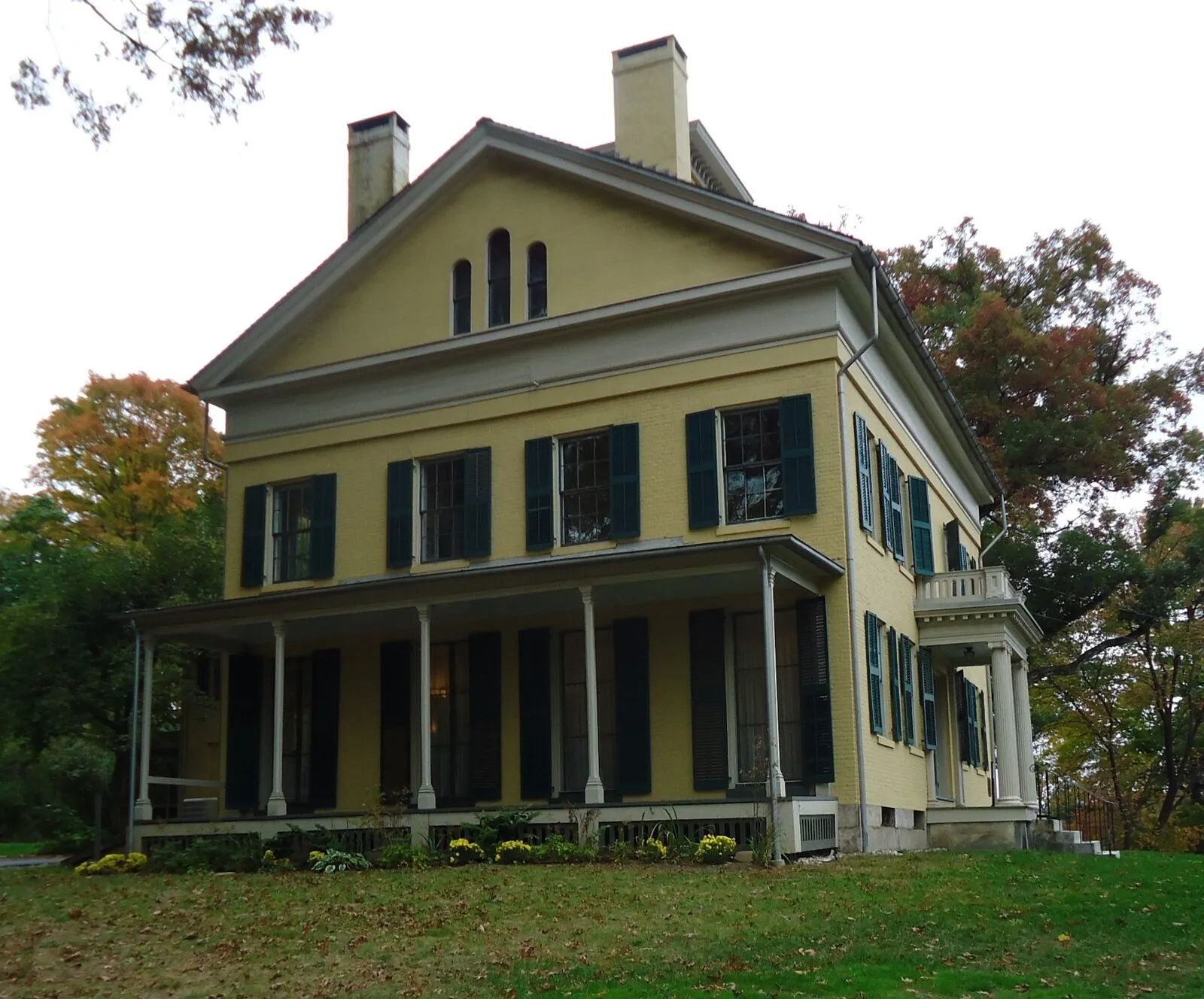 The Emily Dickinson Homestead in Amherst on a clear October afternoon — a two-and-a-half-story Federal-style brick house painted ochre yellow with white trim, three bays on the front with a small portico over the central door, twelve-over-twelve sash windows on both stories and small dormers in the slate-gray roof, a low boxwood hedge in front of the porch, mature deciduous trees in late-fall foliage at left and right framing the house, and a band of clear blue sky above.