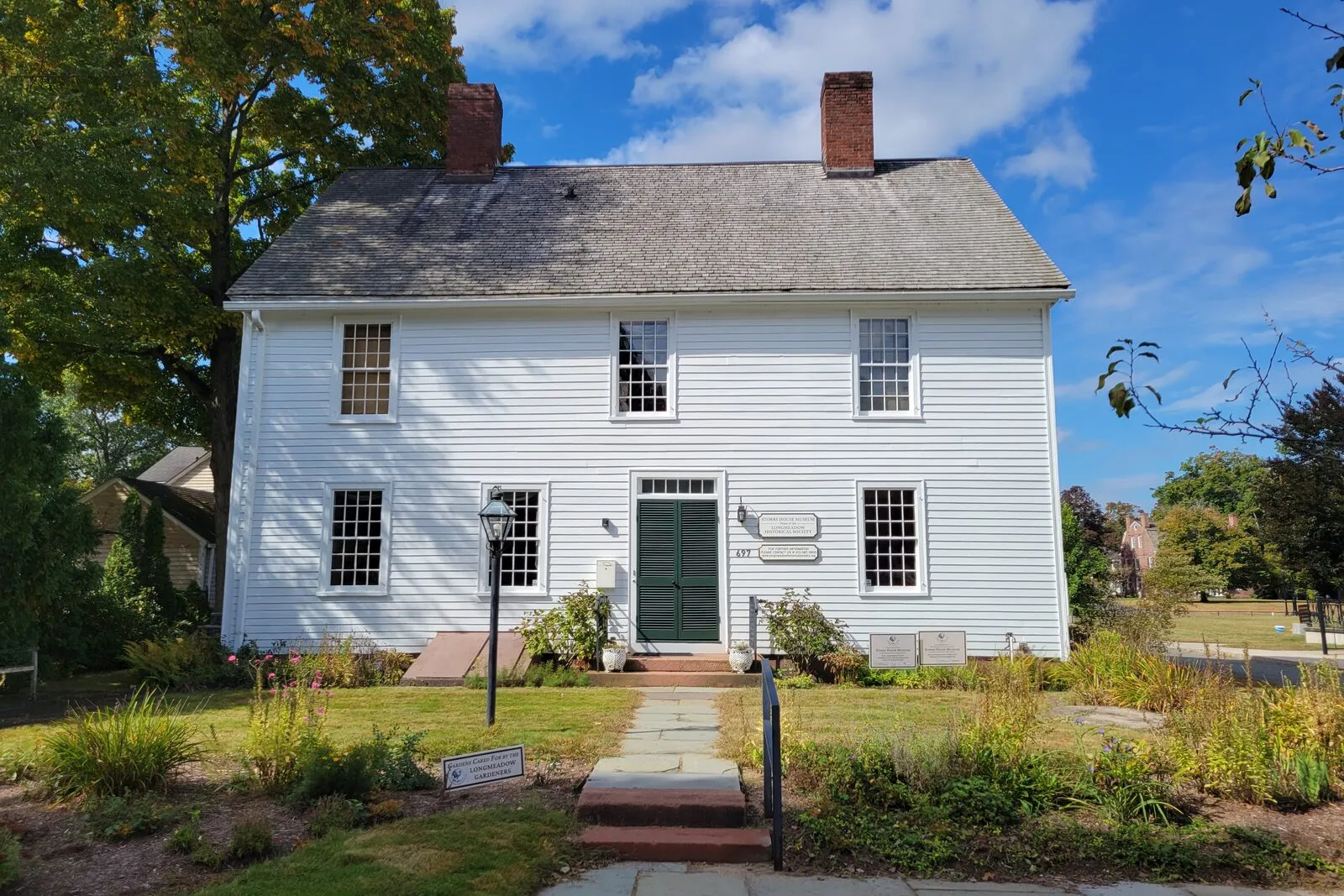 The Storrs House Museum on Longmeadow Street on a sunny early-fall afternoon: a two-and-a-half-story Federal-era white-clapboard center-chimney colonial with a steeply pitched roof, two interior brick chimneys flanking the ridge, five-bay symmetrical façade with twelve-over-twelve sash windows on both stories, a small dormer at the attic, a recessed center entry with a panelled green door under a simple pediment, an exposed brick foundation, a granite-flagged front walk leading up to the door from the sidewalk, perennial-garden borders along both sides of the foundation in late-summer flower, mature deciduous trees in full green leaf framing the house at left and right, and a deep blue sky scattered with thin white clouds overhead.