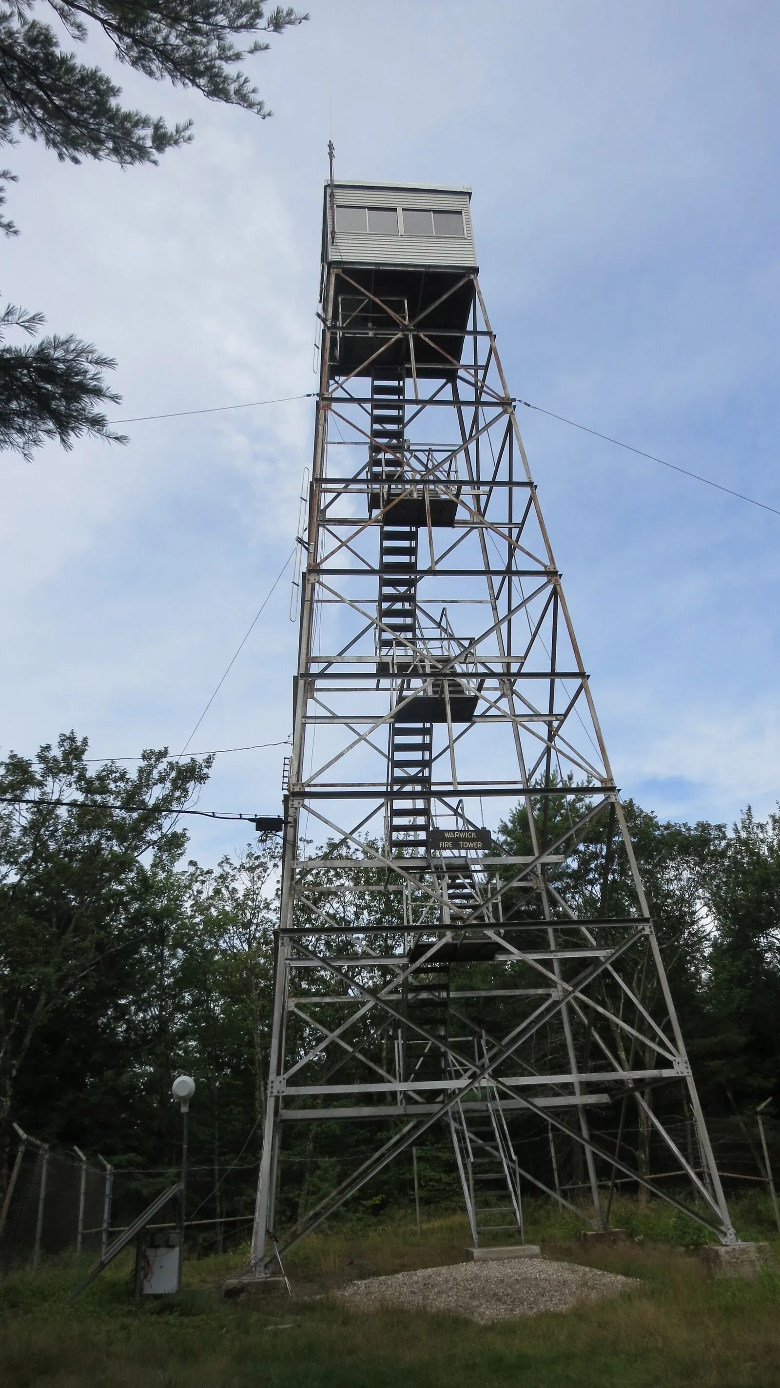 The Warwick Fire Tower on the summit of Mount Grace photographed from the ground looking straight up — a tall, square, four-legged steel lattice tower with a small enclosed wooden cab at the top, several flights of switchback stairs zig-zagging up the inside, the structure rising above a low stone-and-concrete footing on a small clearing of mowed grass and gravel, dark green pines and deciduous trees framing the tower on both sides, and a soft blue summer sky with thin clouds behind.