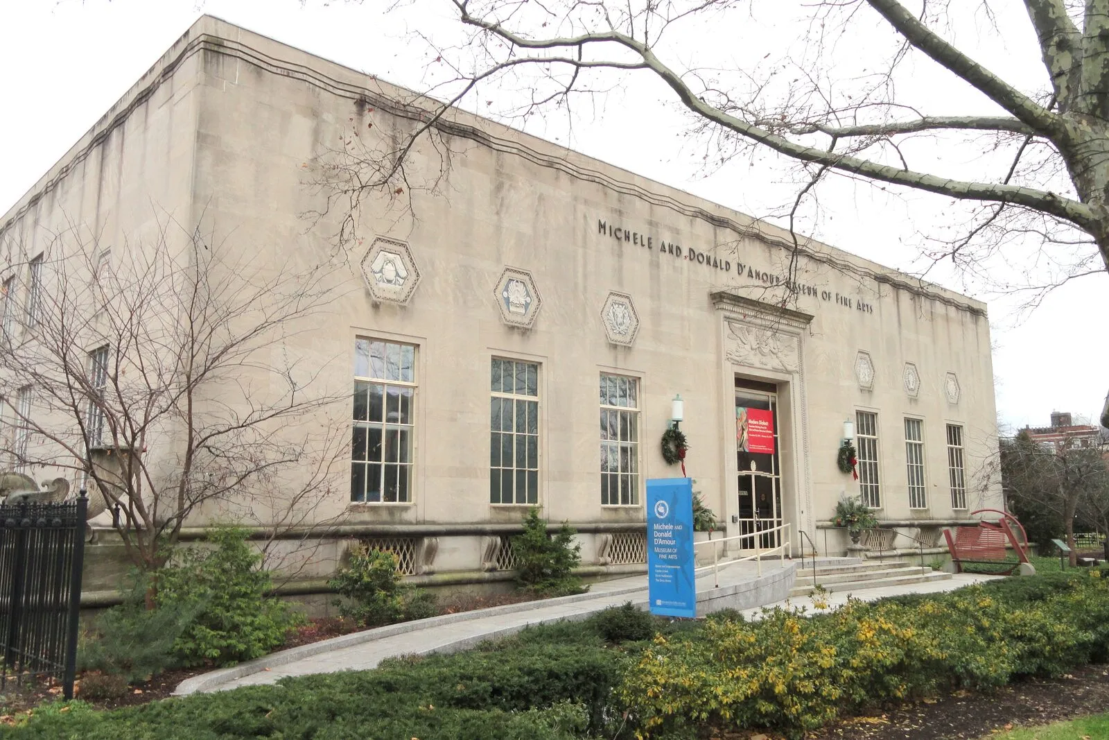 The D'Amour Museum of Fine Arts on Springfield's Quadrangle: a cream-limestone Art Deco museum building with the name carved in a frieze across the façade, octagonal medallion windows along the upper wall, and the central entrance framed by manicured hedges under a gray winter sky.