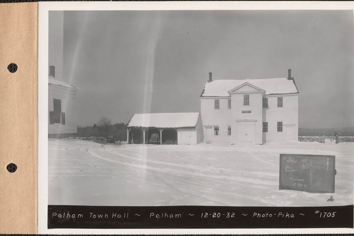 An archival December 1932 black-and-white photograph of the Old Pelham Town Hall in winter. A tall two-story white-clapboard meetinghouse with a low pediment over a single central entry, paired sash windows flanking the door and across the second story, a small open-front shed with a hipped roof off to the left, snow covering the foreground and roofs, a small slate field-tag in the lower right reading 'R-366 / Pelham Town Hall / Pelham 12-20-32', and a hand-lettered banner along the bottom edge of the album page reading 'PELHAM TOWN HALL — PELHAM — 12-20-32 — PHOTO·PIKE — #1705'.