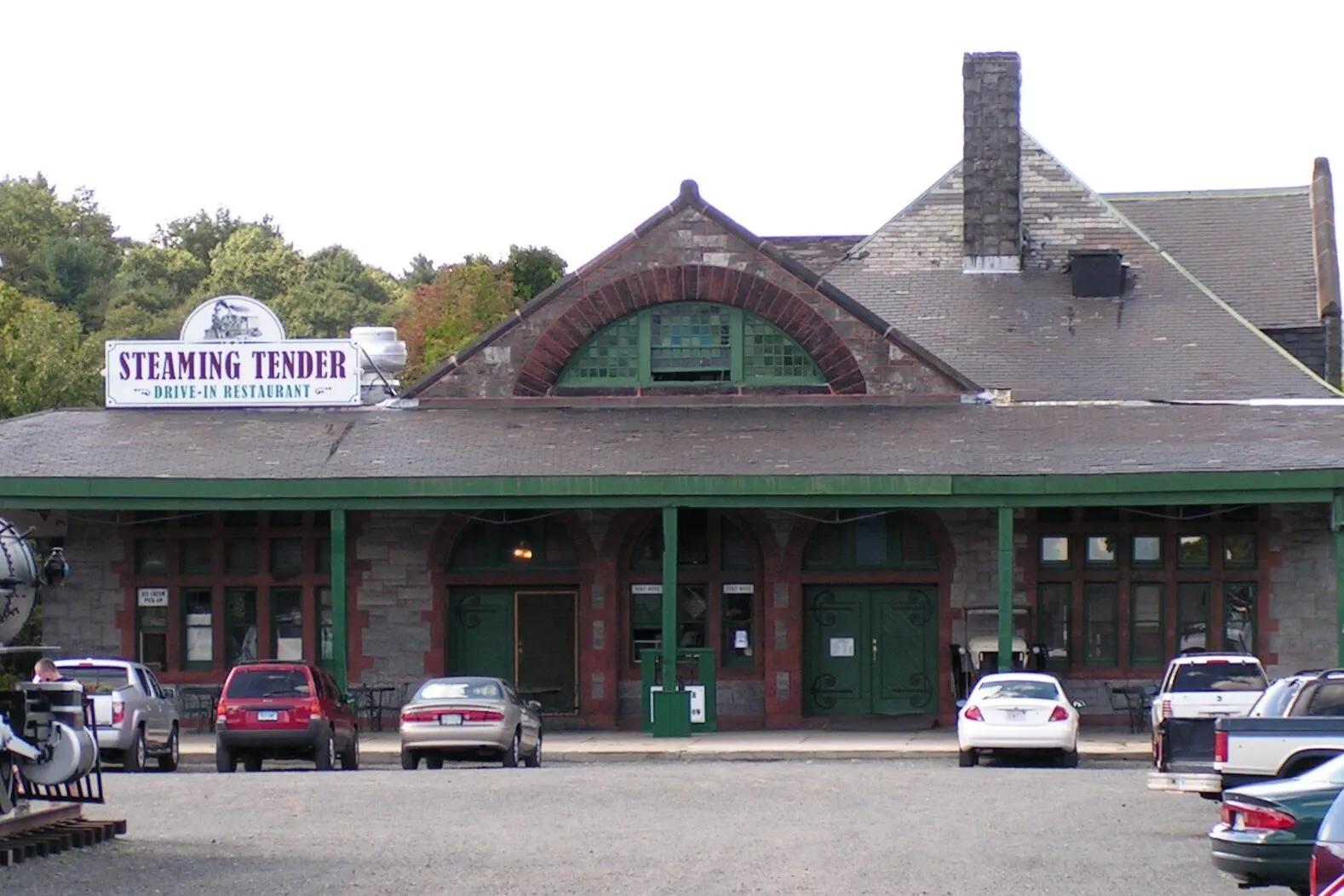 A wide front view of Palmer's historic Union Station. A low, broad H. H. Richardson stone depot with a deep wraparound porch supported by exposed wooden rafters, a steep central gable with a half-round arched window of green-mullioned glass framed in red sandstone, brownstone arches over the platform doors, a tall brick chimney rising on the right, and a small dark steam locomotive parked at the left edge in front of the building. A large white sign above the porch reads STEAMING TENDER DRIVE-IN RESTAURANT. A few late-model cars are parked along the front in the dirt lot, and wooded hillsides rise behind the depot under an overcast sky.