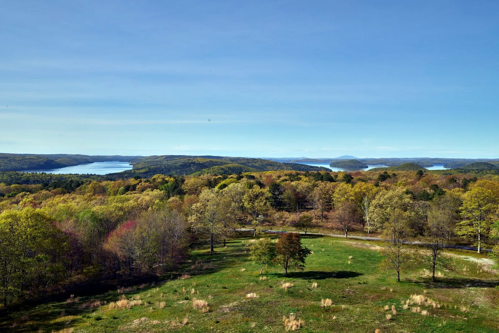View from Quabbin Hill in Quabbin Park on a clear October morning: a wide green grass field at the base of a low hilltop in the foreground, deciduous trees in patchy yellow-and-green fall color filling the middle ground, the long blue ribbons of the Quabbin Reservoir stretching across the middle distance between low forested ridges, more rolling forested hills receding toward the horizon, and a clear pale-blue sky overhead.