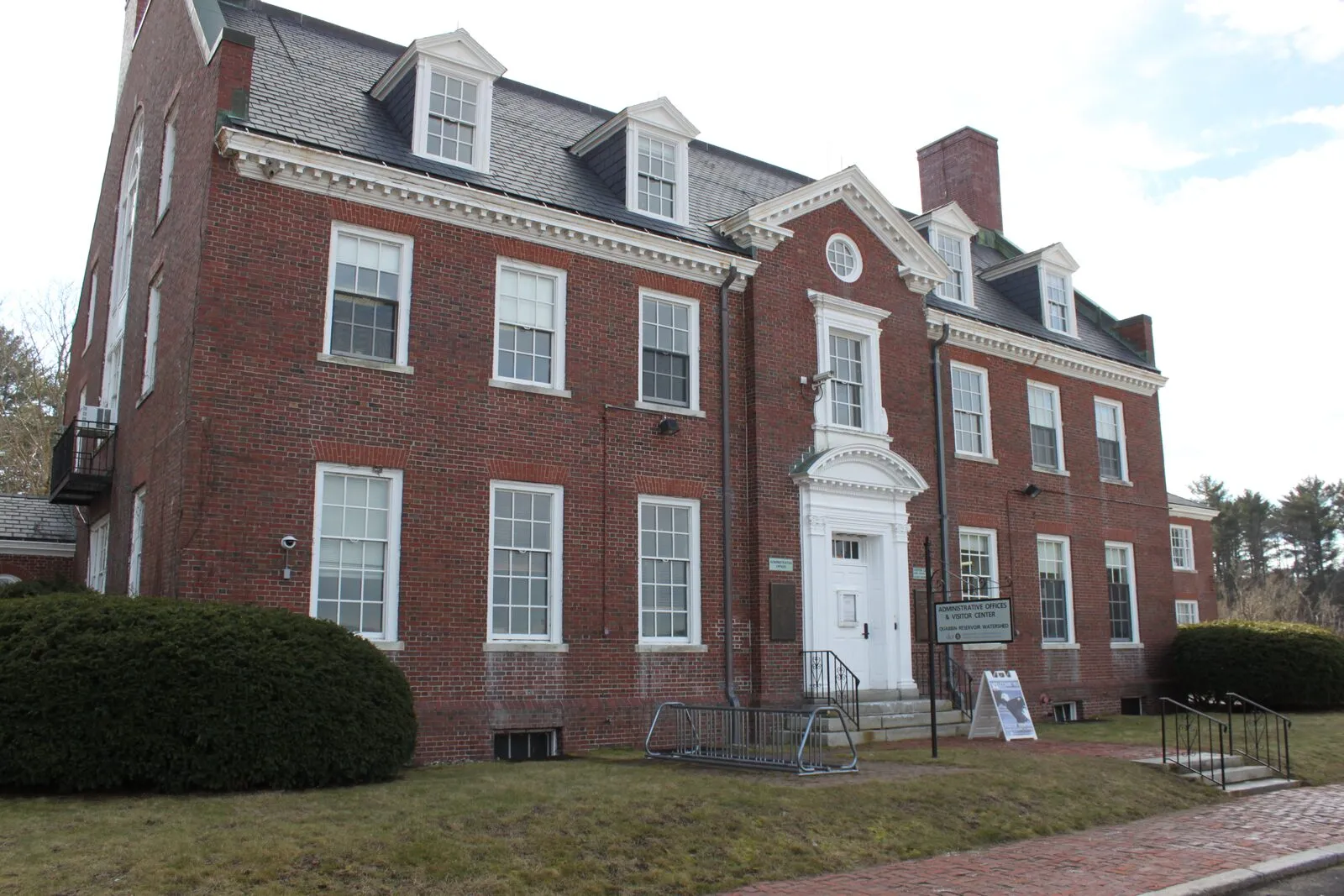 The front elevation of the Quabbin Visitor Center on a clear winter afternoon: a two-and-a-half-story red-brick Federal-style building with a slate roof and three pedimented dormers, two tall brick chimneys, twelve-over-twelve white-trimmed double-hung windows, a central white-columned portico over the front door with a fanlight medallion above, a low boxwood hedge and bare trees on the grass verge in front, a brick walkway leading to the entrance, and a sandwich-board sign reading 'VISITOR CENTER' on the path beside the steps.