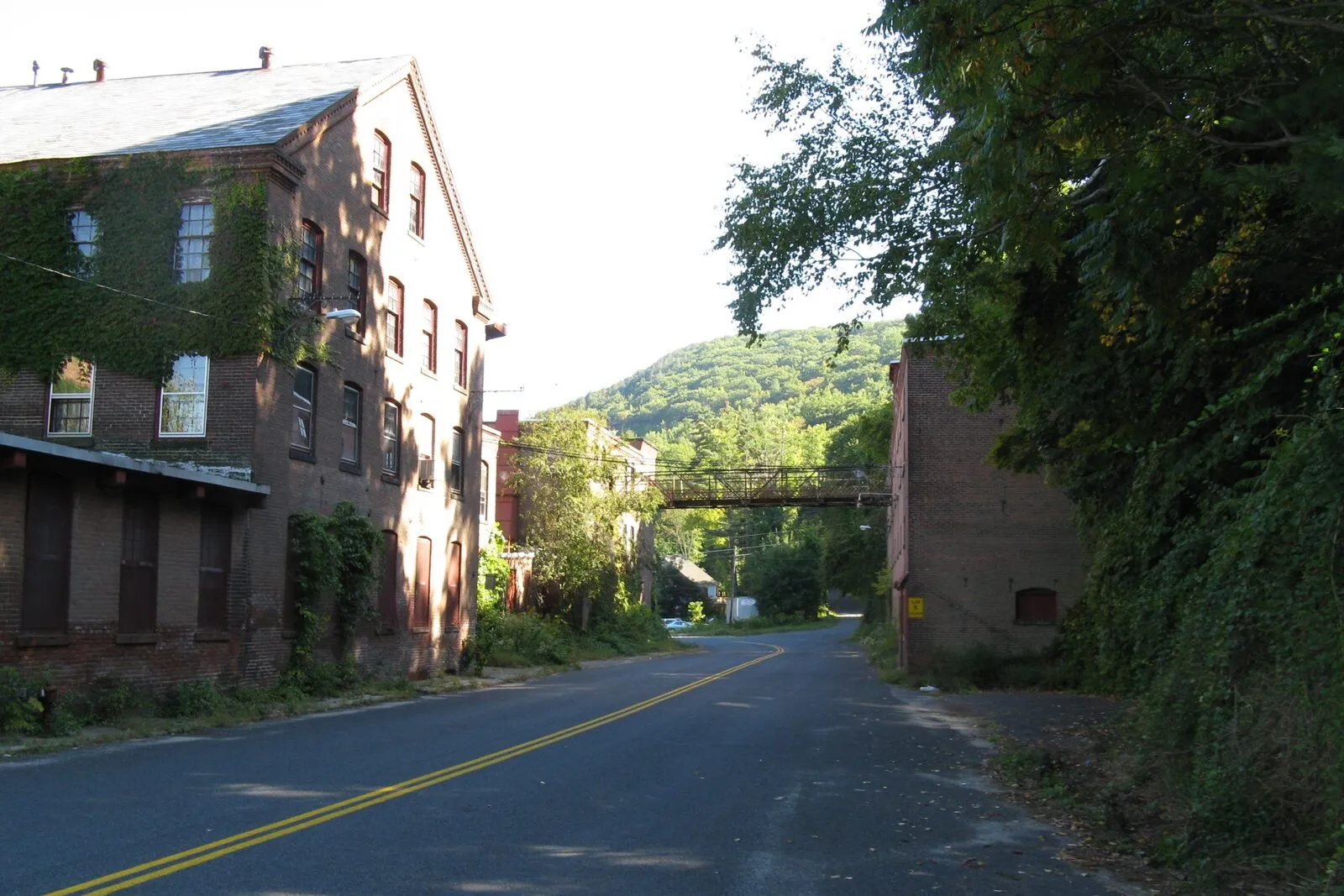 A late-summer afternoon view along Old Route 20 through Woronoco village in Russell: a tall four-story brick paper-mill building of the former Strathmore Mills runs along the left side of a quiet two-lane road, ivy climbing across its lower stories and rows of tall multi-pane factory windows up the wall, a covered overhead skyway crossing the road in the middle distance to a smaller brick mill block on the right, a wooded ridge of the Berkshire foothills rising directly behind the buildings, dense green tree cover spilling in from the right, and a clear pale sky overhead.