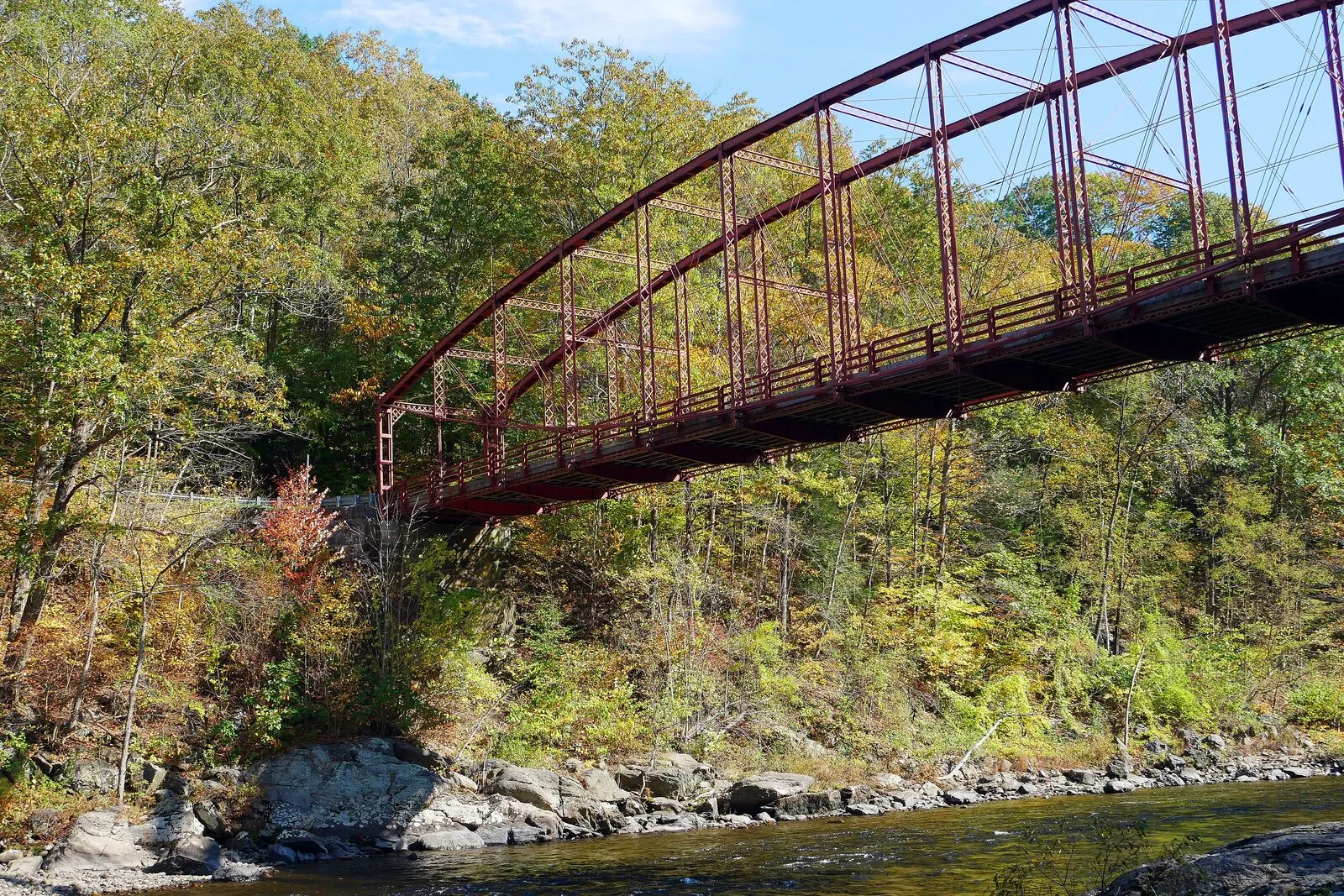 Bardwell's Ferry Bridge from a riverbank angle on a sunny October afternoon — a deep red wrought-iron lenticular-truss bridge curving in a long flattened arch across the Deerfield River, the woods on the far bank in deep yellow and orange fall color, the near bank a tangle of small saplings and shrubs in mixed color, dark gray rock outcrops and a few small boulders along the riverbed in the foreground, the river running clear and shallow over them, and patches of pale blue sky visible through the truss.