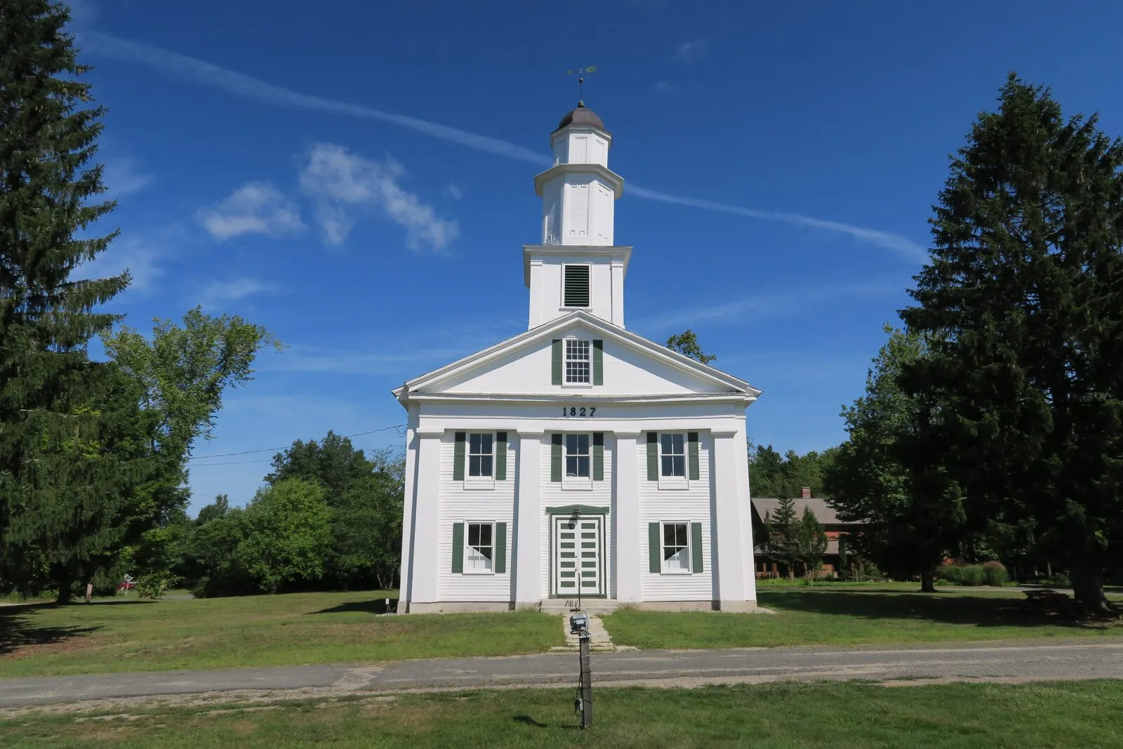 The Shutesbury Community Church on a clear summer afternoon at the village center: a tall white-clapboard Greek Revival meetinghouse with a broad pedimented gable carrying the year 1827 in raised numerals between two flanking second-story windows, a recessed central entry door with louvered shutters, two tiers of large six-over-six sash windows on the front, a lower entablature wrapping the gable, a multi-stage square belfry rising from the ridge with louvered openings and a small dark dome, a weathervane on top, large dark conifers framing the building on both sides, mowed grass and a paved lane in the foreground, and a deep blue sky with thin cirrus clouds overhead.