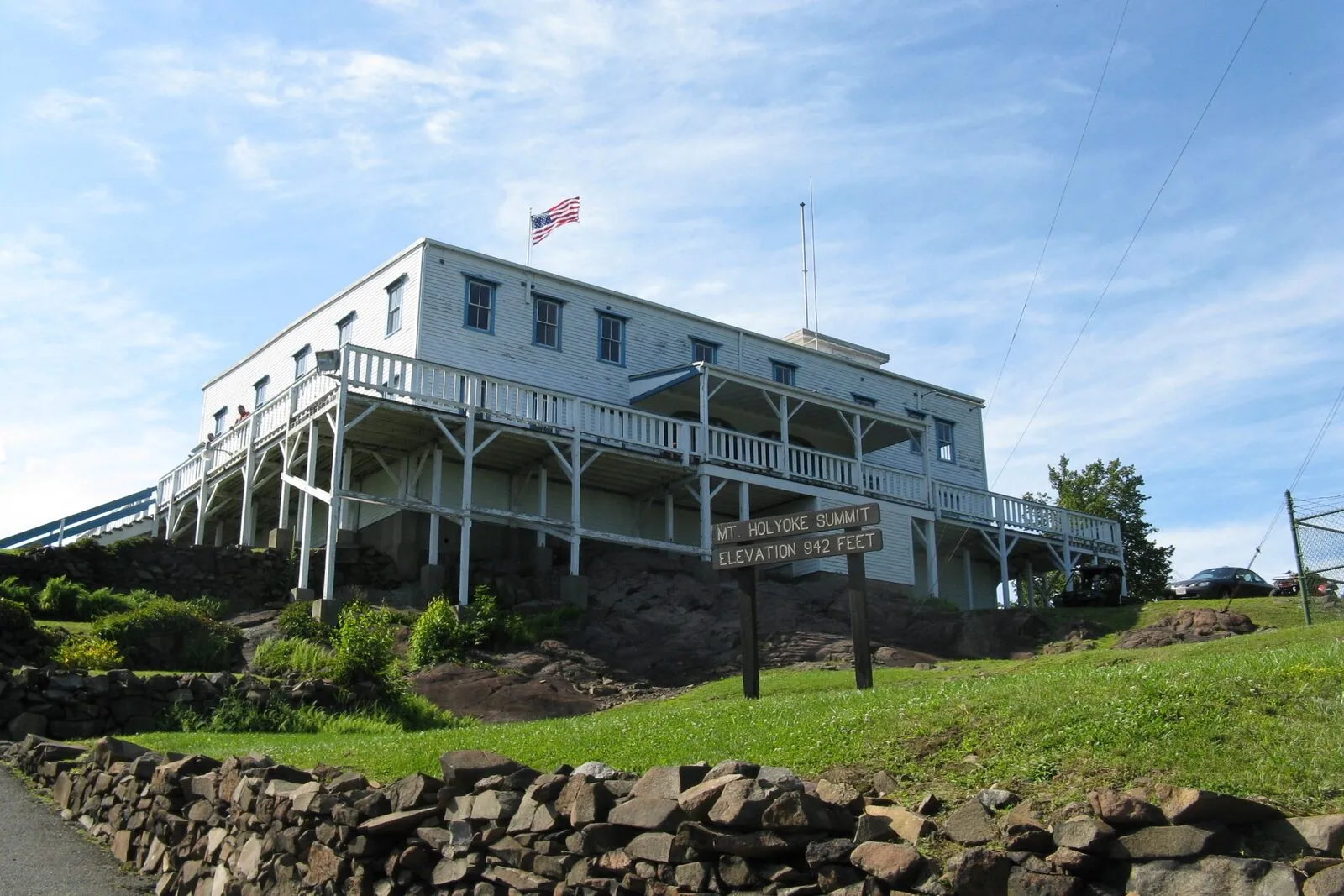 The Summit House at Mount Holyoke, a two-story white clapboard hotel on a stone foundation with a wraparound porch, American flag flying from the roof, and a weathered summit sign in the foreground.