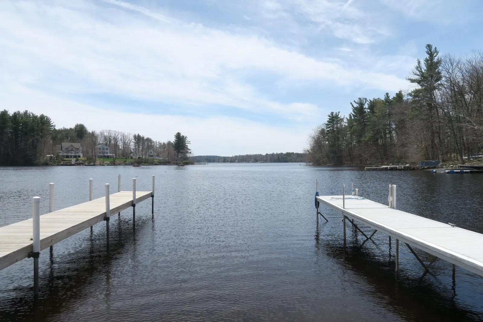 Otis Reservoir on a clear spring afternoon. Two long wooden docks angle out from the foreground into the dark, calm water, framing a wide open view down the lake. A few lakeside houses sit among bare-branched hardwoods on the left shore, a tall stand of hemlock and pine rises on the right shore, and forested hills close the distance under a pale blue sky scattered with thin cirrus clouds. The reservoir is the recreational core of Tolland State Forest.