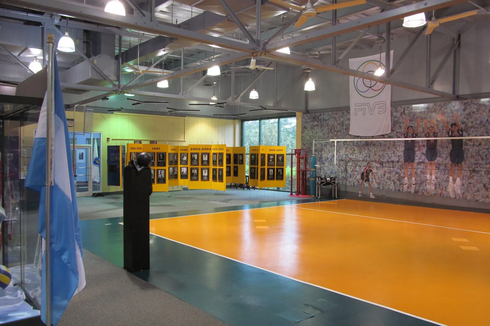 The interior of the International Volleyball Hall of Fame in Holyoke. A regulation-size volleyball court with an orange playing surface and green out-of-bounds borders set inside a converted mill warehouse, a tall white volleyball net stretched across the middle, an FIVB logo and a photo mural of crowd and players on the back wall, banks of yellow-and-gray inductee panels lining the far wall, exposed steel ceiling trusses and pendant lighting overhead, an Argentine flag hanging on the left, and a polished dark green concrete floor reflecting the lights.