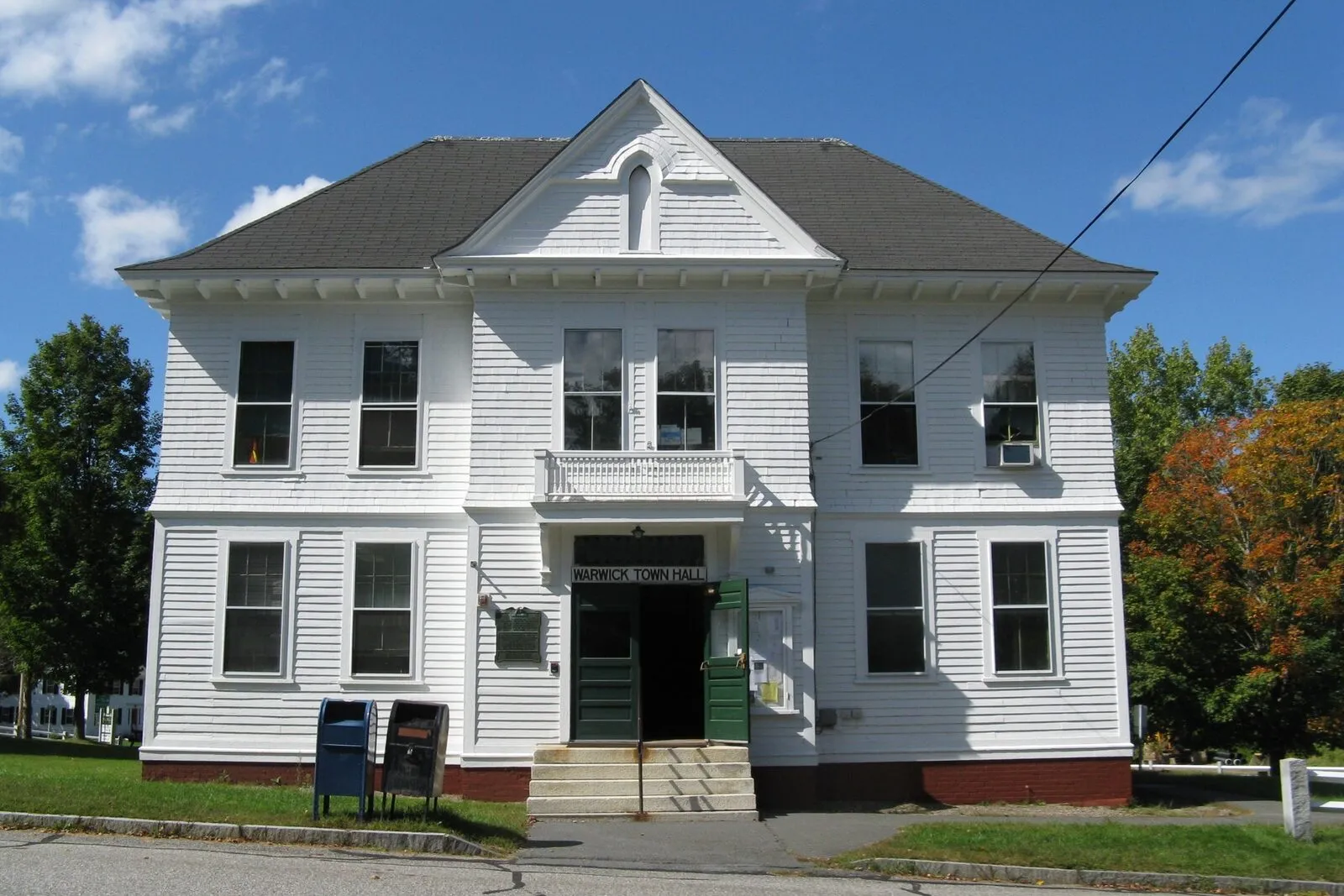 Warwick Town Hall on a clear early-fall afternoon. A two-story white-clapboard 19th-century civic building with a low-pitched hipped roof, a small front-gabled pediment with a round attic vent, two pairs of tall double-hung windows on each side of a centered gabled entry porch, modest wood steps leading up to the porch, a black hand-railing, white-painted wooden front door panels, a blue Postal Service mailbox at the lower-left corner, a clipped green lawn with a low concrete curb in the foreground, dark mature deciduous trees in leaf at right with a few yellowing leaves, a high-tension utility line overhead, and a deep blue sky.