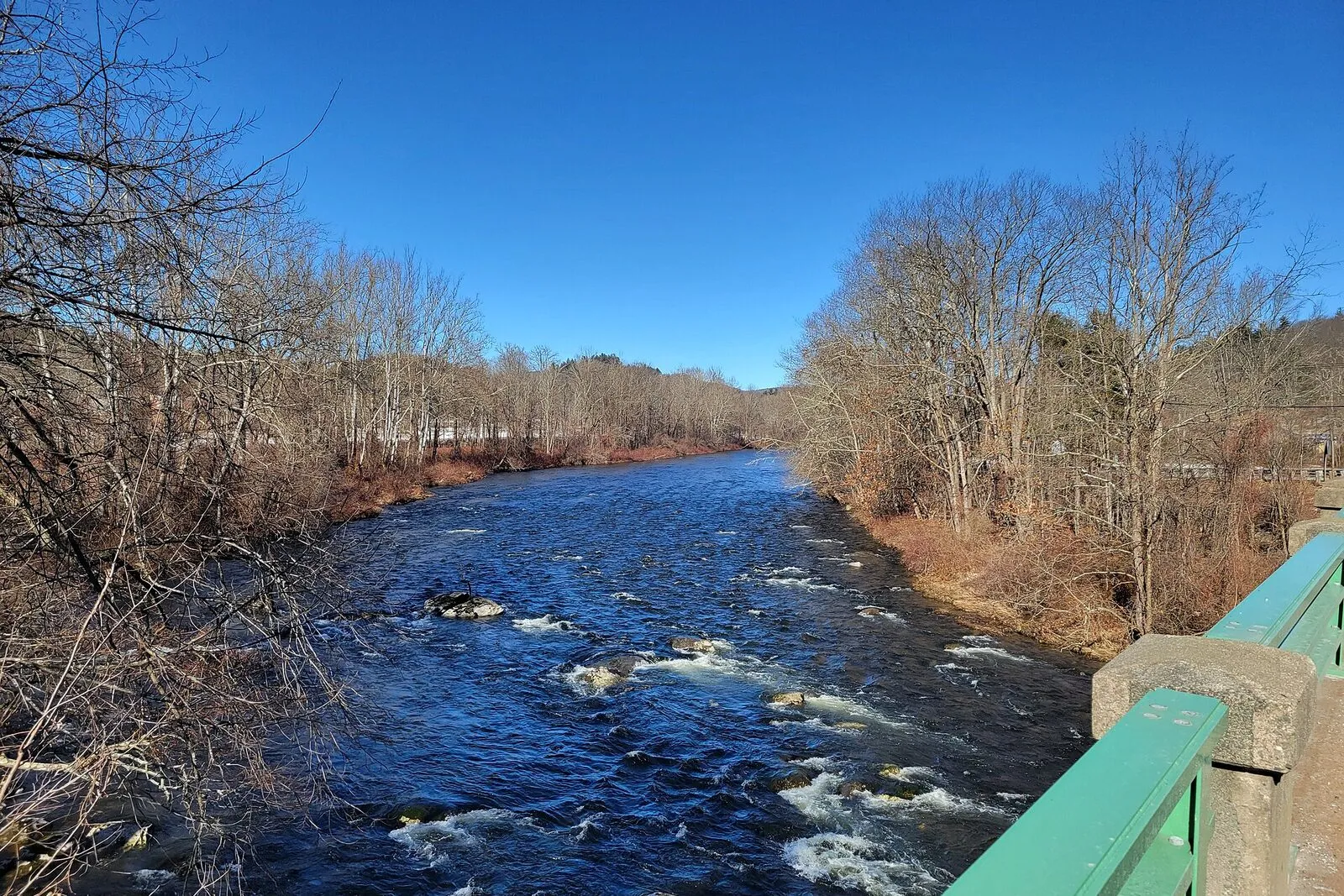 The Westfield River at Norwich Bridge in Huntington on an overcast winter afternoon — a wide, swift stretch of clear river running over a cobble bed scattered with boulders, fast white water breaking through the riffles in the foreground, dense bare-branched leafless deciduous forest on both banks rising up the gorge slopes, a low wooded ridge hemming in the far bank, and a flat gray-white sky overhead.