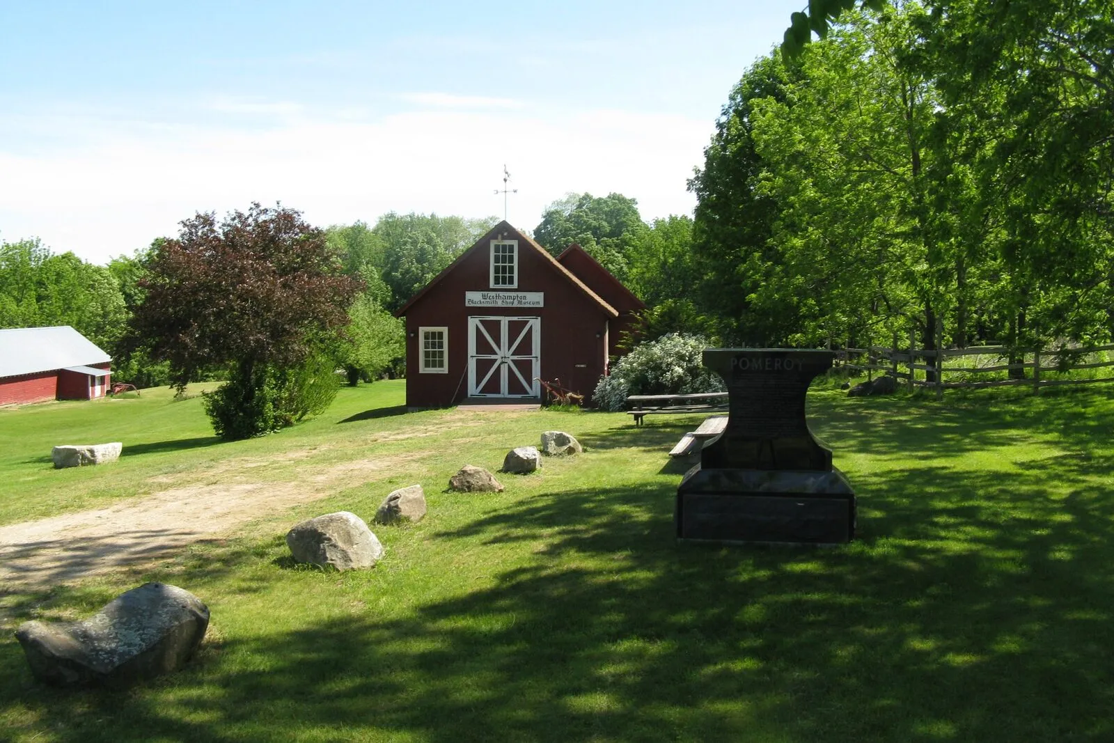 The small Blacksmith Shop Museum on a sunny late-May afternoon in Westhampton: a single-story dark-red clapboard shop with a steeply pitched gable roof, a small dormer over the front, twin white-painted plank doors at the center under a small signboard, a weathervane at the ridge, a heavy black iron anvil and stand on a stone pedestal in the right foreground, low fieldstone boulders scattered across a green mown lawn, a glimpse of a second red outbuilding at the far left, mature deciduous trees in full green leaf framing the scene on both sides, and a clear blue sky overhead.