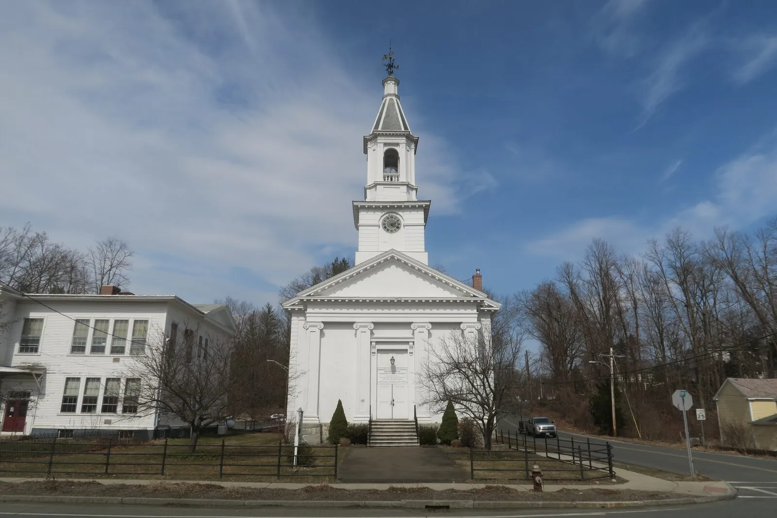 The Haydenville Congregational Church on a clear, breezy late-March afternoon in Haydenville village — a tall white-clapboard Italianate church with a large pedimented gable supported by paired pilasters over a single central entry door, an octagonal bell-stage tower above the gable carrying a clock and a small bell-shaped ogee dome with a weathervane and gilt finial on top, a lower side wing on the left, a small white parsonage barely visible beyond, bare leafless trees flanking on both sides, a wrought-iron fence and bare grass around the foundation, and a deep blue sky streaked with thin cirrus clouds overhead.