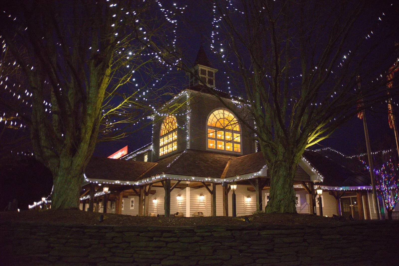The Yankee Candle Village flagship store at night during the holidays. A barn-style building with a central white-railed cupola and an arched fanlight window glowing warm yellow above the entry, white string lights wrapped around the bare branches of mature deciduous trees flanking either side, strands of icicle lights along the eaves, more colored lights on the right wing, and a low fieldstone wall in the foreground under a dark winter sky.