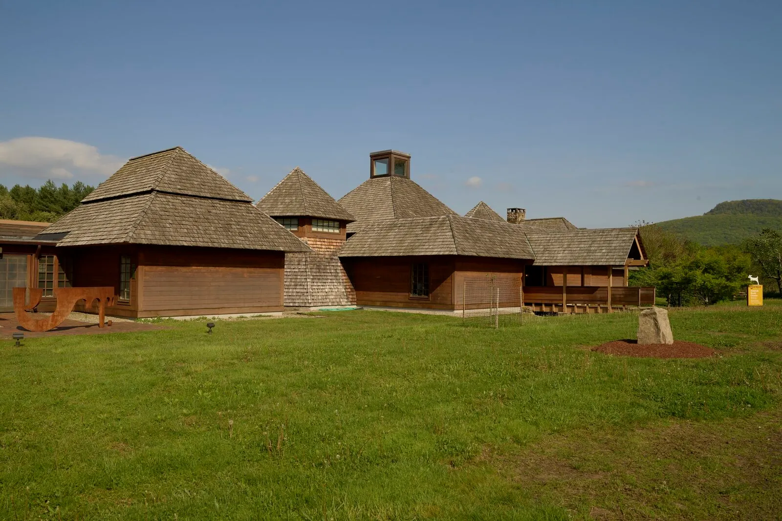 The Yiddish Book Center on a sunny day in late spring, a low cluster of brown wood-shingled buildings with shake-clad pyramidal and conical rooflines arranged across a level green lawn, a forested ridge of the Holyoke Range visible in the far distance on the right under a clear blue sky with thin clouds.