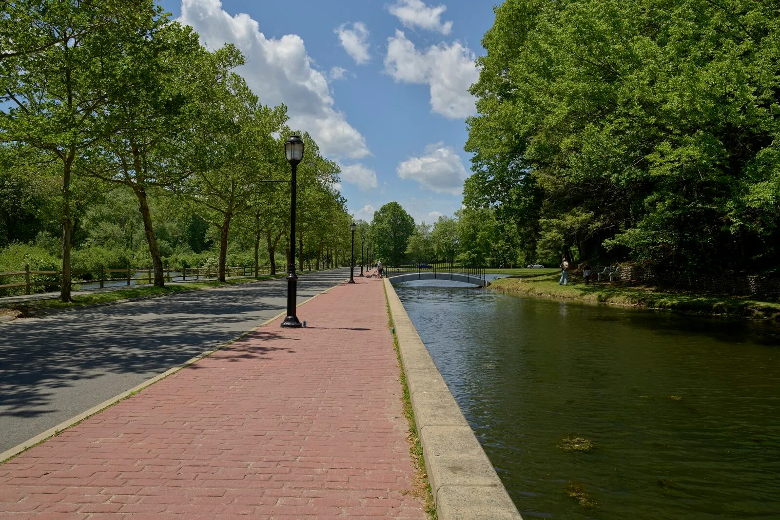The brick-paved walkway alongside a long rectangular pond at Forest Park in Springfield on a sunny late-May morning — classical black cast-iron lampposts spaced along the path, mature broadleaf trees arching over the lawn at left, a low stone-walled embankment edging the water at right, a small arched footbridge spanning the pond in the middle distance, and a bright blue sky with scattered cumulus clouds overhead.